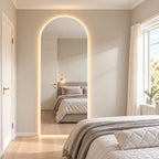 Espejo de pared en arco con luz LED cálida junto a una ventana, con una cama reflejada en un dormitorio de tonos neutros.