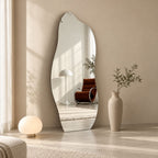 Espejo orgánico de cuerpo entero apoyado en la pared, junto a una lámpara esférica y un florero alto, con un sillón reflejado.