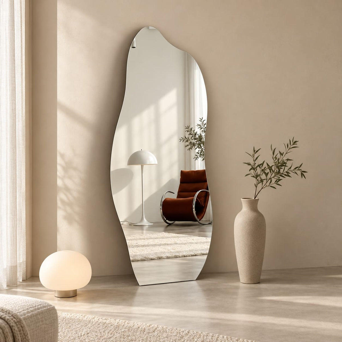 Espejo orgánico de cuerpo entero apoyado en la pared, junto a una lámpara esférica y un florero alto, con un sillón reflejado.