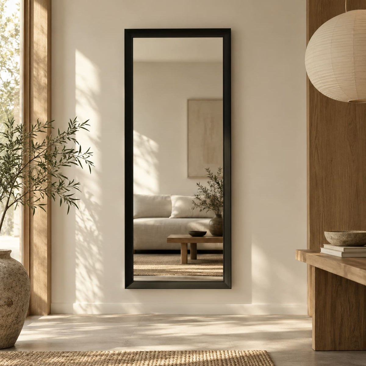 Espejo de pared vertical con marco negro en un recibidor de tonos cálidos, junto a un arrimo de madera y una planta.