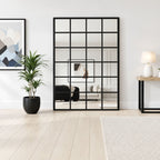 Espejo grande estilo ventana con marco negro y divisiones cuadriculadas, apoyado en el suelo junto a una planta.