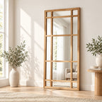 Espejo rectangular estilo ventana con marco de madera natural, apoyado contra una pared junto a un ventanal y un florero